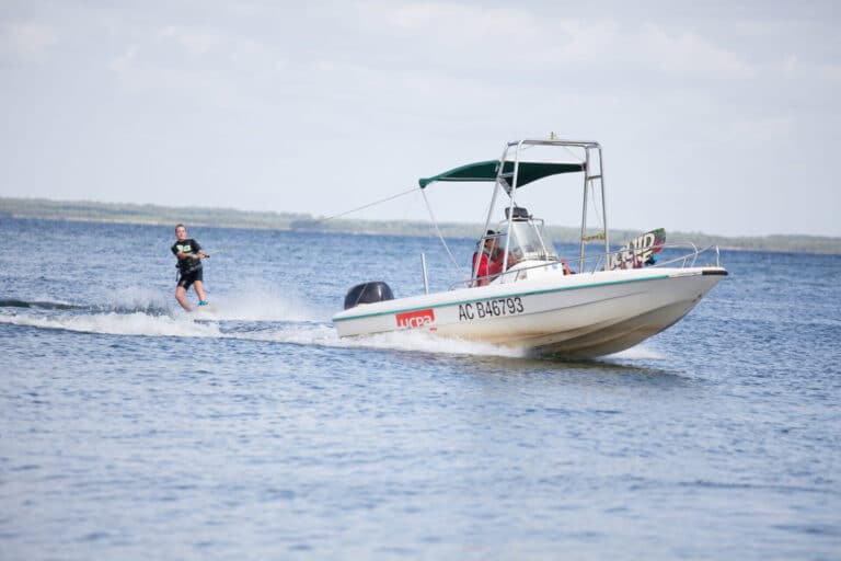 En person wakeboarder på vandet bag en motorbåd med en grøn baldakin. Båden bevæger sig med fart og skaber bølger, og wakeboarderen holder balancen på bølgerne. Baggrunden viser en klar himmel og en fjern kystlinje.