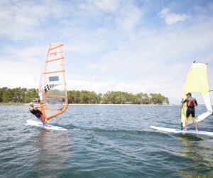 To personer, der windsurfer på et roligt vandområde under en delvist overskyet himmel. Den ene manøvrerer et bræt med et orange sejl, mens den anden har et gult sejl. Træer langs den fjerne kystlinje.