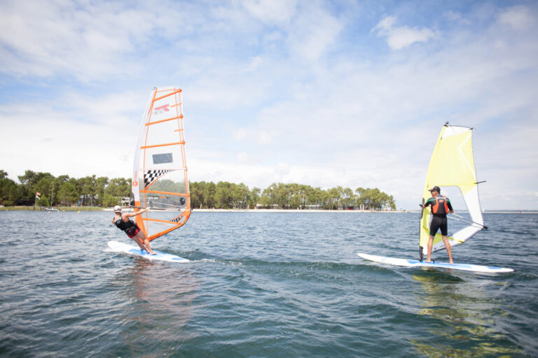 To personer, der windsurfer på et roligt vandområde under en delvist overskyet himmel. Den ene manøvrerer et bræt med et orange sejl, mens den anden har et gult sejl. Træer langs den fjerne kystlinje.