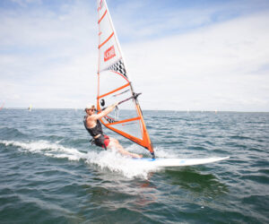 Person, der windsurfer på et stort vandområde under en klar blå himmel, iført redningsvest og boardshorts. Sejlet er orange med ternet mønster. Bølger bølger i vandet, og andre windsurfingsejl er synlige i det fjerne.