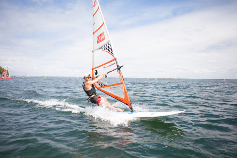 Person, der windsurfer på et stort vandområde under en klar blå himmel, iført redningsvest og boardshorts. Sejlet er orange med ternet mønster. Bølger bølger i vandet, og andre windsurfingsejl er synlige i det fjerne.