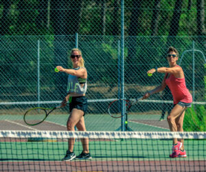 To personer på en udendørs tennisbane, begge holder ketchere og forbereder sig på at tjene. De er iført sportsudstyr, inklusive shorts og ærmeløse toppe, med et grønt hegn og træer i baggrunden.