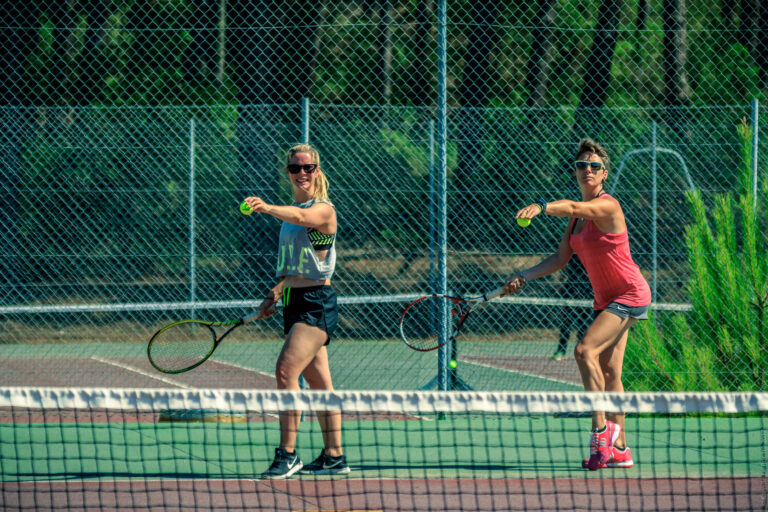 To personer på en udendørs tennisbane, begge holder ketchere og forbereder sig på at tjene. De er iført sportsudstyr, inklusive shorts og ærmeløse toppe, med et grønt hegn og træer i baggrunden.