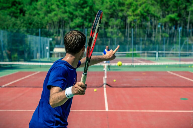 En tennisspiller i en blå skjorte forbereder sig på at slå en bold på en lerbane med en skovklædt kulisse. En anden spiller står i den modsatte ende af banen, klar til at modtage skuddet.