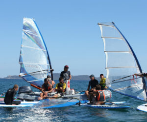 En gruppe mennesker på windsurfboards og en lille båd er samlet på en solskinsdag på havet. De er iført afslappet vandsportstøj, og den klare blå himmel er i baggrunden.