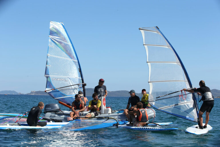 En gruppe mennesker på windsurfboards og en lille båd er samlet på en solskinsdag på havet. De er iført afslappet vandsportstøj, og den klare blå himmel er i baggrunden.
