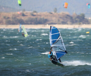 En windsurfer rider på bølgerne på en blæsende dag med et blåt sejl, omgivet af andre windsurfere i det fjerne. Baggrunden har et kuperet landskab under en klar himmel, og havet er hakket med hvide huer.