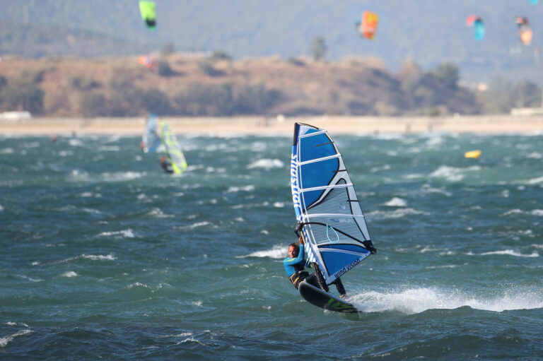 En windsurfer rider på bølgerne på en blæsende dag med et blåt sejl, omgivet af andre windsurfere i det fjerne. Baggrunden har et kuperet landskab under en klar himmel, og havet er hakket med hvide huer.