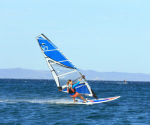 En person, der windsurferer på havet under en klar blå himmel, med fjerne bakker synlige i horisonten. De er iført en orange redningsvest og manøvrerer et blåt og hvidt sejl.