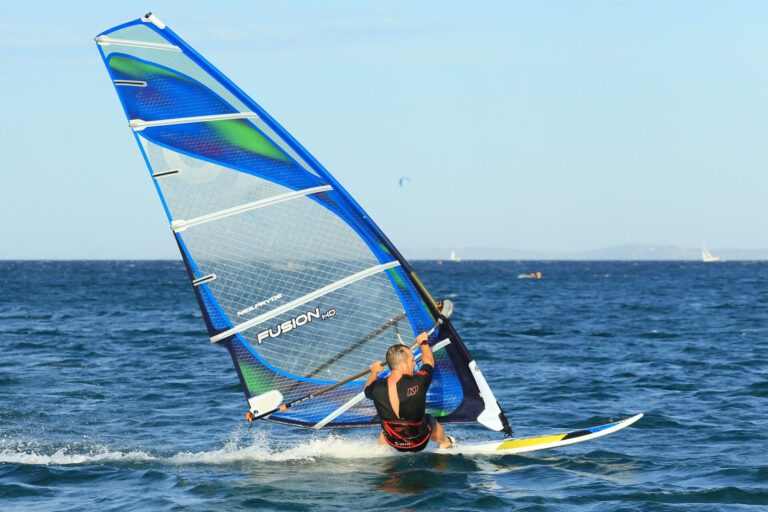Person windsurfing på havet med et blåt og grønt sejl. Windsurferen er vinklet over brættet, mens de glider over vandet. Klar blå himmel og fjern horisont i baggrunden.