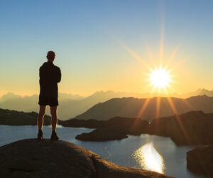 En person står på en klippe, silhuet mod en levende solnedgang over et bjergrigt landskab. Sollyset reflekteres fra en rolig sø med fjerne tinder synlige i horisonten. Scenen formidler en følelse af ro og fordybelse.