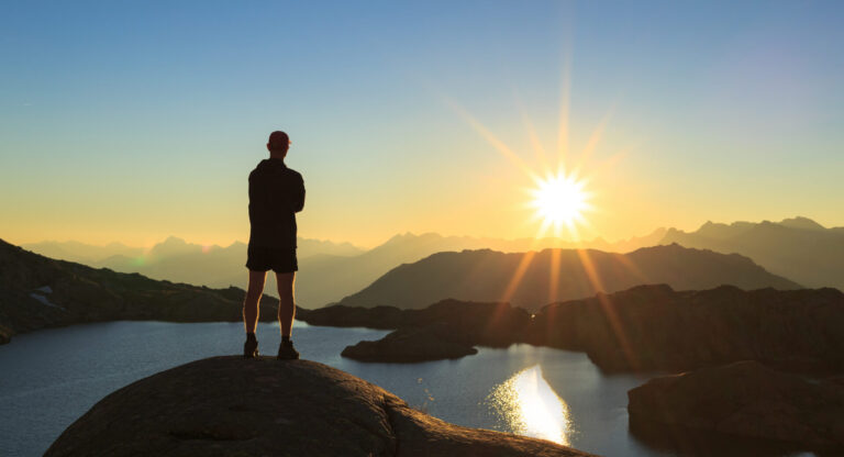 En person står på en klippe, silhuet mod en levende solnedgang over et bjergrigt landskab. Sollyset reflekteres fra en rolig sø med fjerne tinder synlige i horisonten. Scenen formidler en følelse af ro og fordybelse.