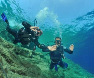 To dykkere under vandet i klart blåt vand, iført sorte våddragter og dykkerudstyr. De svømmer over en stenet havbund. En dykker giver et OK håndsignal, og begge ser ud til at nyde dykket.