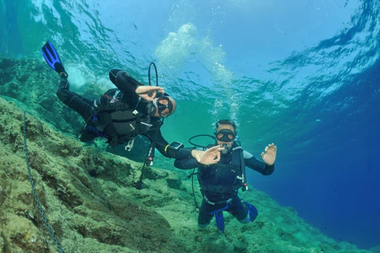 To dykkere under vandet i klart blåt vand, iført sorte våddragter og dykkerudstyr. De svømmer over en stenet havbund. En dykker giver et OK håndsignal, og begge ser ud til at nyde dykket.