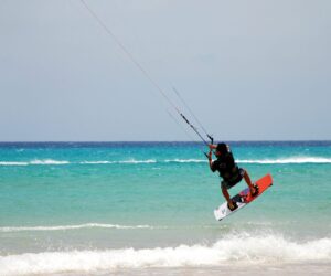 En person kiteboarder over havet og udfører et hop. Det turkise vand står i kontrast til den klare blå himmel. Bølger slår blidt ned nedenfor og skildrer en solrig, eventyrlig dag på stranden.