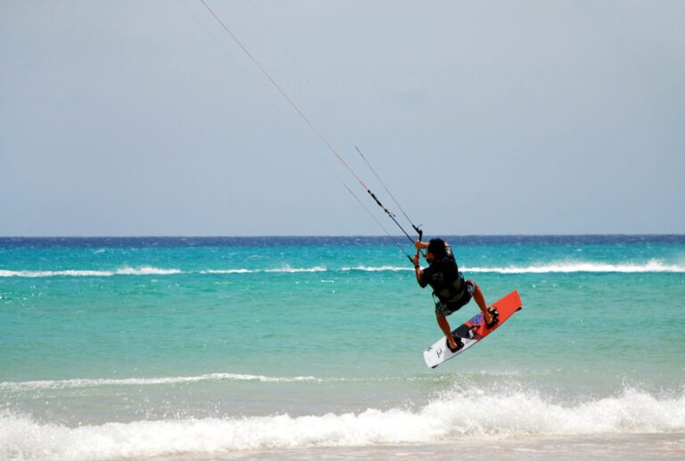 En person kiteboarder over havet og udfører et hop. Det turkise vand står i kontrast til den klare blå himmel. Bølger slår blidt ned nedenfor og skildrer en solrig, eventyrlig dag på stranden.
