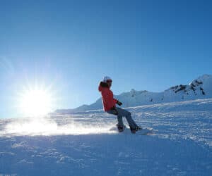 En person snowboarder ned ad en sneklædt bjergskråning på en solskinsdag, hvor solen skinner klart på den klare blå himmel. Sneklædte tinder er synlige i baggrunden.