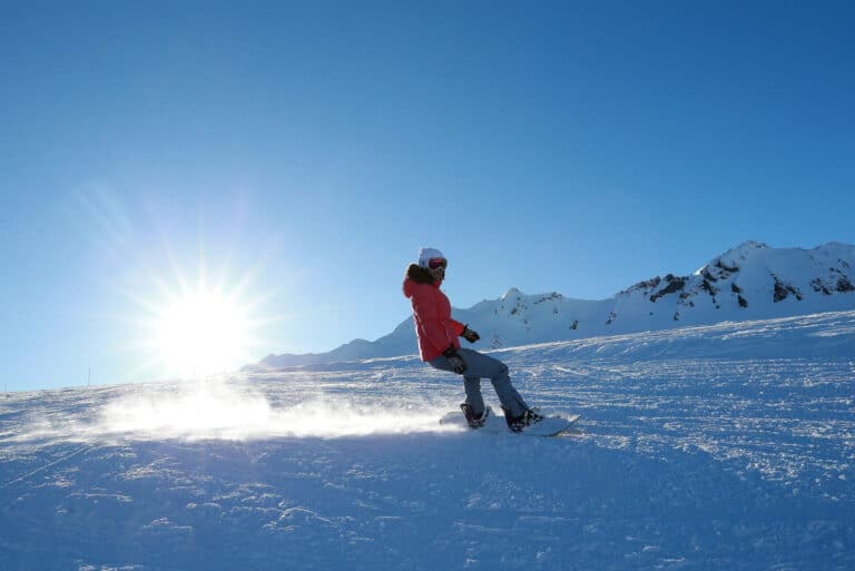 En person snowboarder ned ad en sneklædt bjergskråning på en solskinsdag, hvor solen skinner klart på den klare blå himmel. Sneklædte tinder er synlige i baggrunden.