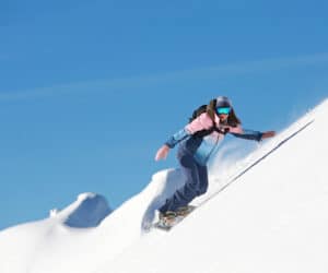 En person snowboarder ned ad en stejl, sneklædt skråning under en klar blå himmel. De bærer en farverig jakke, mørke bukser, reflekterende briller og en hjelm. Sne bliver sparket op omkring dem, mens de navigerer på den uberørte hvide overflade.