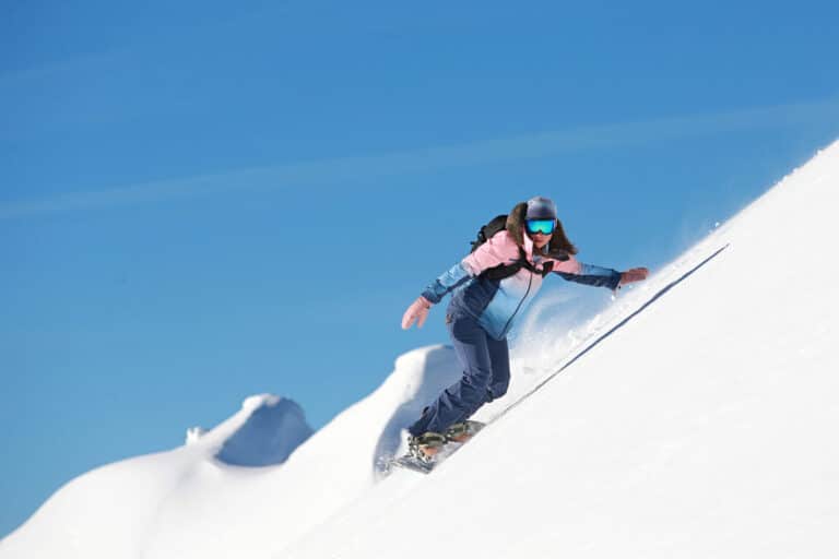 En person snowboarder ned ad en stejl, sneklædt skråning under en klar blå himmel. De bærer en farverig jakke, mørke bukser, reflekterende briller og en hjelm. Sne bliver sparket op omkring dem, mens de navigerer på den uberørte hvide overflade.