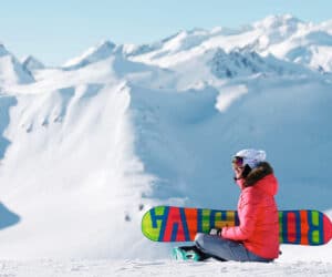 En person i en lys rød jakke og hvid hjelm sidder på en snedækket bjergskråning med et farverigt snowboard. Snedækkede tinder og en klar blå himmel er i baggrunden, hvilket skaber en fredfyldt alpine scene.