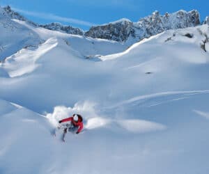 En skiløber i en rød jakke stiger yndefuldt ned ad en sneklædt bjergskråning og skaber et spor af pudder. Baggrunden viser robuste, snedækkede tinder under en klar blå himmel.