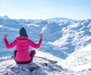 En person i en lyserød jakke og blå hue sidder med krydsede ben på et snedækket bjerg og mediterer. De har udsigt over et stort, solbelyst landskab med snedækkede tinder under en klar blå himmel.