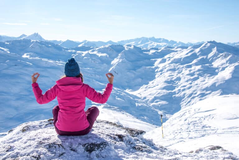 En person i en lyserød jakke og blå hue sidder med krydsede ben på et snedækket bjerg og mediterer. De har udsigt over et stort, solbelyst landskab med snedækkede tinder under en klar blå himmel.