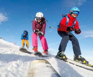 Tre personer står på ski ned ad en sneklædt skråning under en klar blå himmel. De er iført farverige skitøj og hjelme og nyder vinteraktiviteten. Sneen bliver synligt sparket op af deres ski, mens de kører ned ad skråningen.