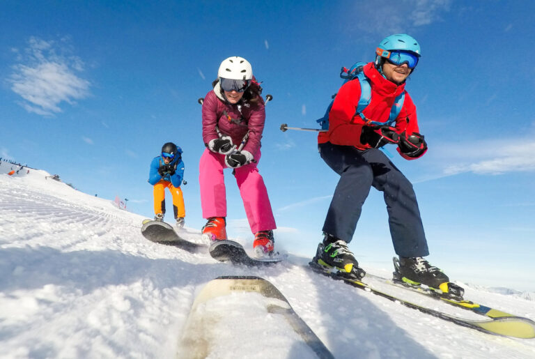 Tre skiløbere i farverigt gear står på ski ned ad bakke på en sneklædt skråning under en klar blå himmel. Den forreste skiløber er i rød og blå, efterfulgt af en skiløber i pink og hvid, og en anden i blå og orange. De nyder den solrige dag.