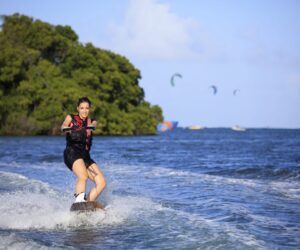 En person kører på et wakeboard over en blå havbugt og holder fast i et reb. Iført redningsvest og shorts karrer de sig gennem vandet med træer og kitesurfere synlige i baggrunden under en klar himmel, og inkarnerer spændingen ved en eventyrlig sportsferie.