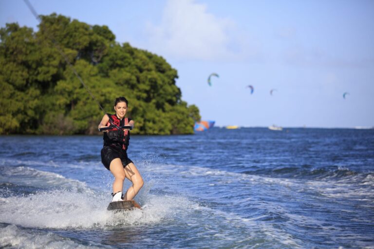 En person kører på et wakeboard over en blå havbugt og holder fast i et reb. Iført redningsvest og shorts karrer de sig gennem vandet med træer og kitesurfere synlige i baggrunden under en klar himmel, og inkarnerer spændingen ved en eventyrlig sportsferie.