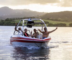 Tre personer på en lille motorbåd smiler og gestikulerer energisk, som om de deler historier om deres seneste skiferieeventyr. Båden sejler gennem stille vand med bakker og en overskyet himmel i baggrunden.