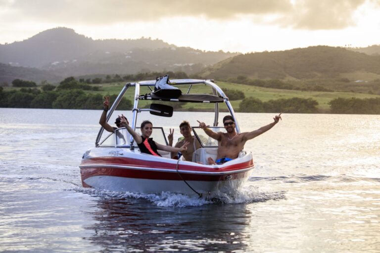 Tre personer på en lille motorbåd smiler og gestikulerer energisk, som om de deler historier om deres seneste skiferieeventyr. Båden sejler gennem stille vand med bakker og en overskyet himmel i baggrunden.