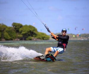 En person er på kiteboarding på en solskinsdag. De er iført en blå hjelm, solbriller og en våddragt og glider over vandet med træer og andre kiteboardere synlige i baggrunden.