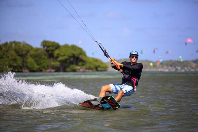 En person er på kiteboarding på en solskinsdag. De er iført en blå hjelm, solbriller og en våddragt og glider over vandet med træer og andre kiteboardere synlige i baggrunden.