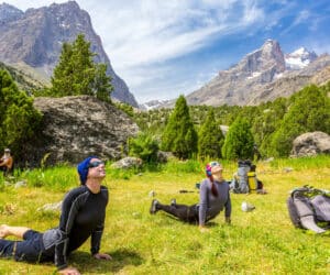 To mennesker dyrker yoga i en frodig bjergdal under en blå himmel. De er på et græsklædt område omgivet af træer og klipper med høje sneklædte tinder i baggrunden. Rygsække hviler i nærheden.