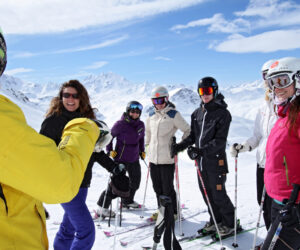 En gruppe skiløbere klædt i farverigt vintertøj står med ski på en sneklædt bjergskråning. De smiler og sludrer under en klar blå himmel med fjerne snedækkede tinder i baggrunden.