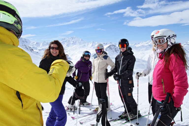 En gruppe skiløbere klædt i farverigt vintertøj står med ski på en sneklædt bjergskråning. De smiler og sludrer under en klar blå himmel med fjerne snedækkede tinder i baggrunden.
