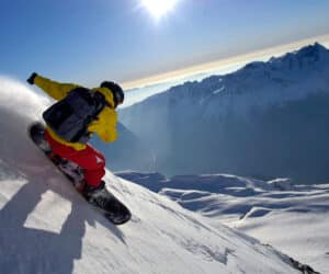 En snowboarder iført en gul jakke og røde bukser går ned ad en snedækket bjergskråning. Solen skinner klart på den blå himmel, med forrevne bjergtoppe synlige i baggrunden. Snesprøjter bag snowboardet.
