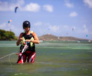 En person iført hjelm og solbriller står på lavt vand og styrer kiteboarding-udstyr. I baggrunden flyver flere drager på himlen på baggrund af fjerne bakker og spredte skyer.
