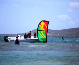 Folk samles på en båd i havet nær et farverigt kiteboard. En person er på kiteboarding i det fjerne. Baggrunden har en kuperet kystlinje under en blå himmel.