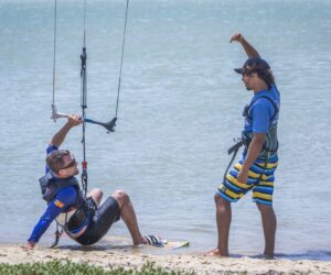 En person sidder på stranden og holder en kiteboarding-kontrolstang, mens en anden person står ved siden af dem og gestikulerer, muligvis giver instruktioner. Begge er iført seler og afslappet strandtøj. Vandet er roligt i baggrunden.