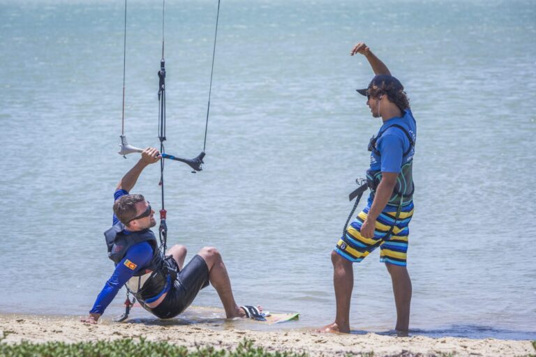 En person sidder på stranden og holder en kiteboarding-kontrolstang, mens en anden person står ved siden af dem og gestikulerer, muligvis giver instruktioner. Begge er iført seler og afslappet strandtøj. Vandet er roligt i baggrunden.