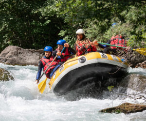 Tre personer iført hjelme og redningsveste navigerer en gul oppustelig tømmerflåde gennem strømfald. De holder pagajer og virker fokuserede. Det omkringliggende område er frodigt med grønne områder og klipper.
