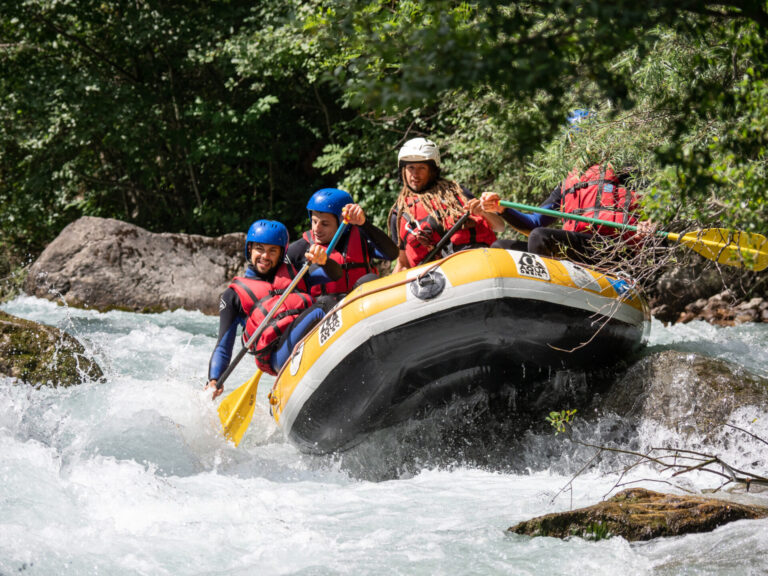 Tre personer iført hjelme og redningsveste navigerer en gul oppustelig tømmerflåde gennem strømfald. De holder pagajer og virker fokuserede. Det omkringliggende område er frodigt med grønne områder og klipper.