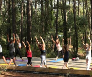 En gruppe mennesker dyrker yoga udendørs i skovmiljøer. De står på yogamåtter, med løftede arme, omgivet af høje træer og grønt. Scenen antyder et roligt, naturligt miljø for motion.
