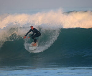 En surfer i en sort våddragt rider på en stor bølge på et surfbræt. Havet er turbulent med hvidt skum, og himlen er let overskyet, hvilket kaster et blødt lys over scenen.