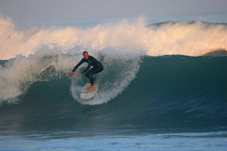 En surfer i en sort våddragt rider på en stor bølge på et surfbræt. Havet er turbulent med hvidt skum, og himlen er let overskyet, hvilket kaster et blødt lys over scenen.