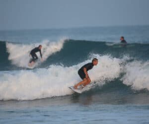 Tre surfere rider på havets bølger. Forgrundssurferen sidder på hug på en bølge midt i bevægelse. En anden surfer er synlig på en bølge i baggrunden, med en tredje i det fjerne. Havet er roligt ud over bølgerne, under en klar himmel.